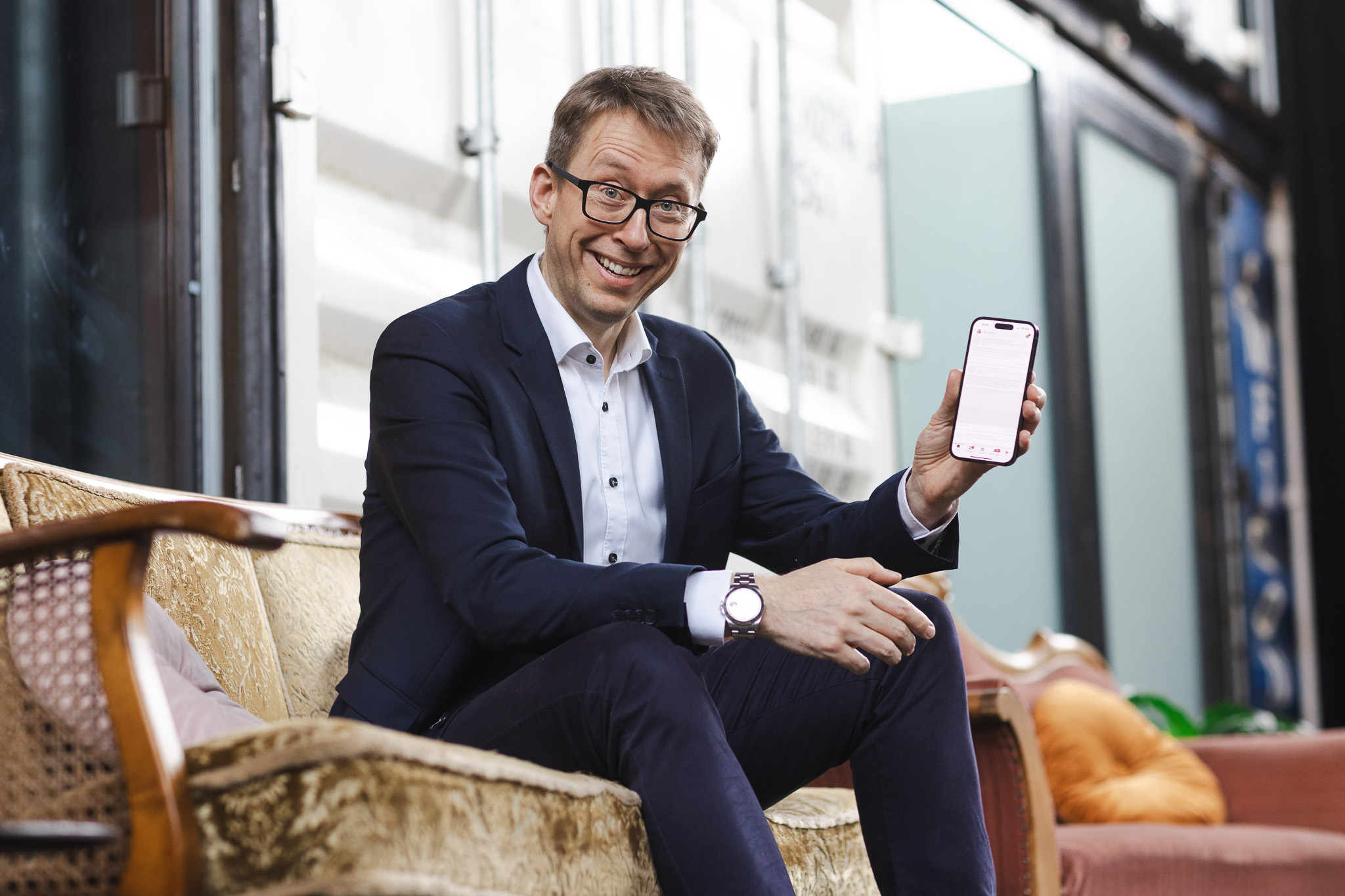 Daniel Schwenger sitzt auf einem Sofa und zeigt lächelnd sein Smartphone